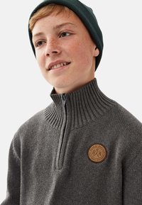 Grå strikket pullovertrøje med en høj ribbet krave og en kvart lynlås. Har et rundt lædermærke på brystet.