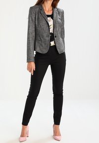 Grå tekstureret blazer med enkelt knaplukning, kombineret med sorte tætsiddende bukser og pink højhælede sko. Blomstret mønstret top nedenunder.