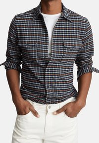 Rutigt skjorta med blå, vita och bruna nyanser, knappar framtill, två bröstfickor och uppvände ärmar. Matchas med vita byxor.