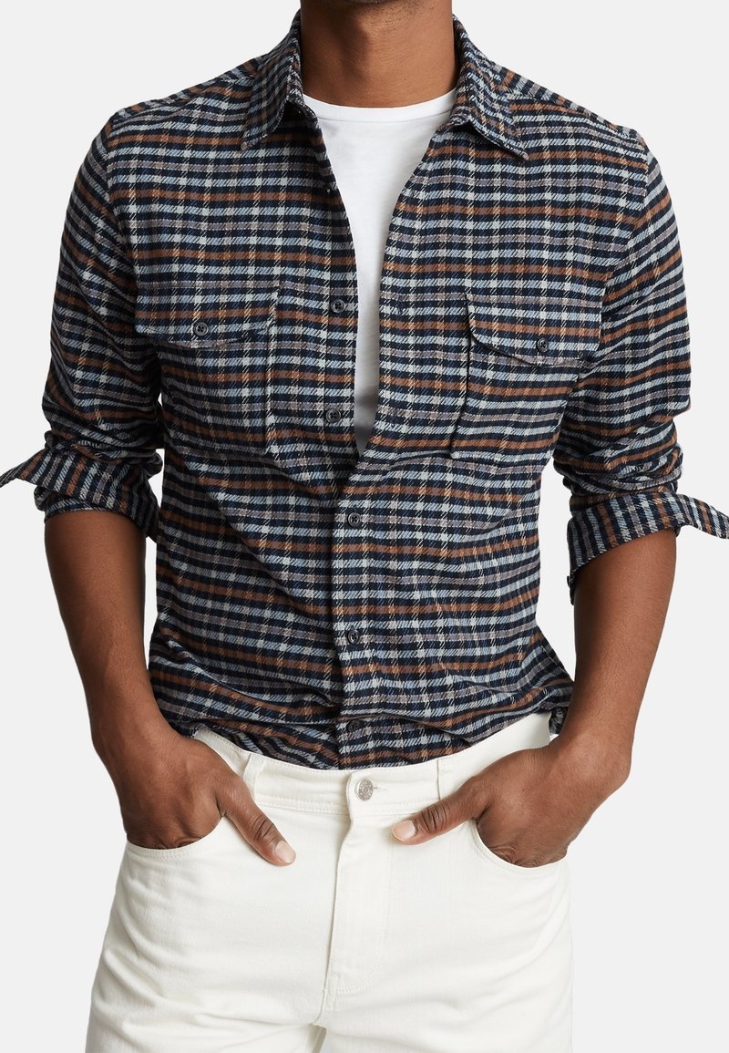 Rutigt skjorta med blå, vita och bruna nyanser, knappar framtill, två bröstfickor och uppvände ärmar. Matchas med vita byxor.