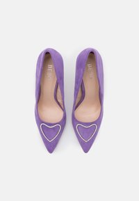 Escarpins en suédine violette avec des pointes aiguës, présentant des découpes en forme de cœur soulignées de strass. L'intérieur est de couleur beige clair.