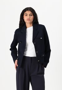Veste bleu marine au design structuré, avec des boutons argentés, deux poches avant et un col cranté. Portée sur une chemise blanche.