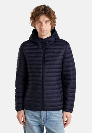 Mężczyzna o kręconych włosach, ubrany w granatową pikowaną kurtkę z kapturem na suwak oraz jasnoniebieskie dżinsy, stojący na jednolitym tle.