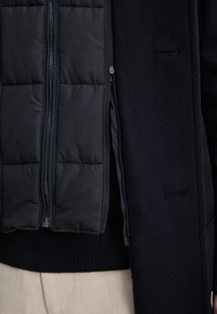 Navy Puffer-Weste mit gestepptem Muster, ausgestattet mit einem Reißverschluss. Darüber ist ein dunkler Wollmantel zu sehen; sichtbarer gerippter Strickbund und Taschendetails.