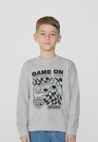 Šedá mikina s dlouhými rukávy, která má černou grafiku herního ovladače a text. Měkká textura a volný střih. Nosená s modrými džíny.