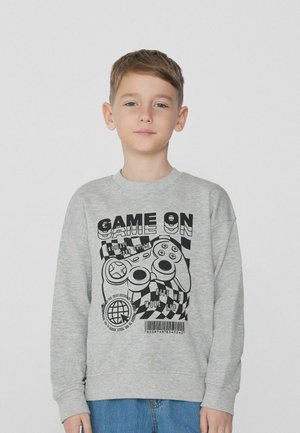 Felpa grigia con maniche lunghe, caratterizzata da una grafica nera di un controller di gioco e testo. Tessuto morbido e vestibilità casual. Indossata con jeans in denim blu.