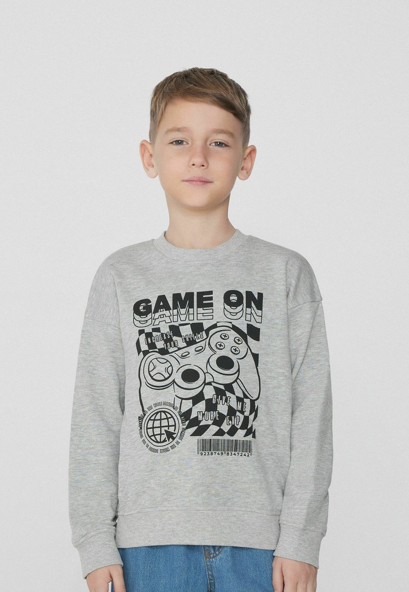 Šedá mikina s dlouhými rukávy, která má černou grafiku herního ovladače a text. Měkká textura a volný střih. Nosená s modrými džíny.