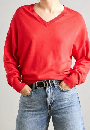 Personne portant un pull à col en V rouge vif rentré dans un jean bleu clair taille haute avec une ceinture noire, mains dans les poches.