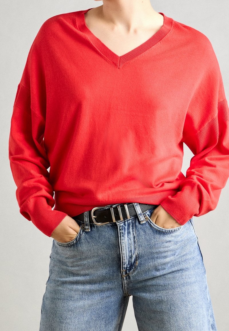 Personne portant un pull à col en V rouge vif rentré dans un jean bleu clair taille haute avec une ceinture noire, mains dans les poches.