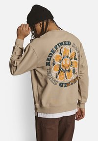 Beige sweatshirt med et stort illustreret blomsterdesign og tekst på ryggen, med lange ærmer og løs pasform. Båret over en hvid skjorte.