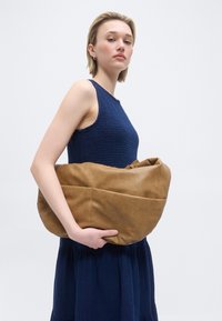 Νεαρή γυναίκα με αμάνικο φόρεμα σε ναυτικό μπλε, κρατώντας μια μεγάλη, μαλακή καφέ δερμάτινη τσάντα μπροστά σε απλό ανοιχτόχρωμο φόντο.