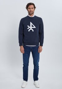 Sudadera azul marino con diseño gráfico blanco centrado en el pecho, combinada con vaqueros azules y zapatillas blancas. Estilo sencillo y casual.