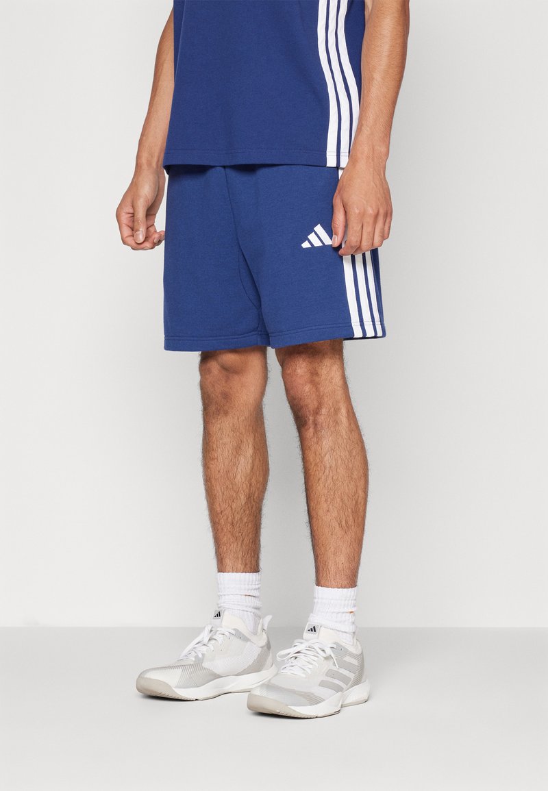 Adidas blauwe sportshorts met witte streepaccenten, gemaakt van een zachte stof, gecombineerd met witte sokken en lichtgrijze sneakers.