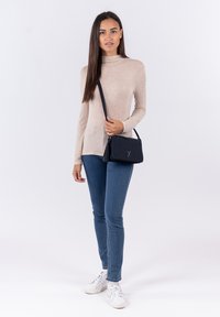 Borsa a tracolla in pelle nera con dettaglio a frange e logo in rilievo, abbinata a una maglietta beige a maniche lunghe e jeans skinny blu.