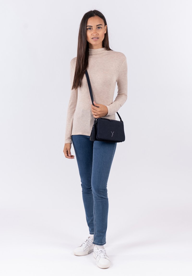 Borsa a tracolla in pelle nera con dettaglio a frange e logo in rilievo, abbinata a una maglietta beige a maniche lunghe e jeans skinny blu.