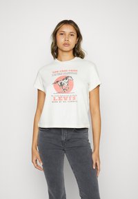 Hvit crop t-skjorte med en rød grafikk av en hest og tekst. Korte ermer og en avslappet passform, kombinert med grå denimjeans.