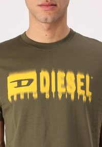 Olivenfarget bomulls t-skjorte med et livlig gult grafisk trykk med ordene "DIESEL" og "DENIM DIVISION," designet med rund hals.