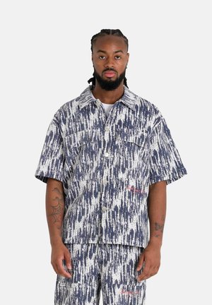 Chemise à manches courtes en tissu texturé avec un motif abstrait bleu marine et blanc, dotée de deux poches avant et de boutons argentés.