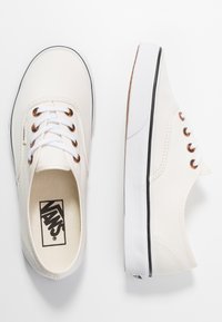 Weiße Leinenschuhe mit runder Spitze, schwarzem Gummisohle und Metallösen; ausgestattet mit weißen Schnürsenkeln und sichtbar im Inneren das Vans-Logo.