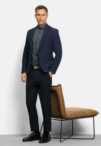 Marineblazer mit slim fit, gemustertem dunklem Hemd, dunkler Hose und schwarzen Schnürschuhen. Die Textur umfasst glattes Material und weiche Stuhlpolsterung.