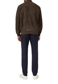 Braune Wildleder-Bomberjacke mit gerippten Bündchen, getragen über marineblauen Hosen mit elastischen Bündchen und weißen Sneakers. Die Rückansicht zeigt den strukturierten Stoff.
