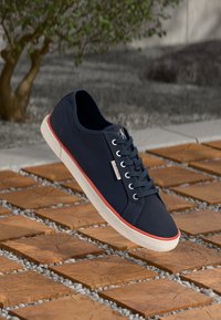 Navy blue canvas sneakers with rounded toe, white rubber sole, red accent stripe, and simple lace-up design. Placed on textured clay tiles.