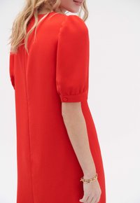 Vestido rojo con mangas cortas abullonadas y detalles de botones en los puños. Textura suave, corte recto, detalles de costura visibles y un perfil minimalista.