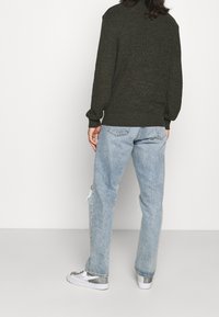 Tmavě zelený svetr s texturovaným pletením a žebrovaným lemem, v kombinaci se světle modrými džínami s potrhaným efektem a šedými teniskami s síťovanými detaily.