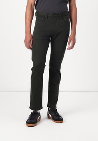 Pantalones ajustados de color verde oscuro, hechos de una tela suave, que cuentan con un diseño clásico de cinco bolsillos y cierre de botón metálico. Combinados con zapatillas negras.