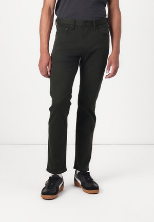 Sötétzöld, slim fit nadrág puha anyagból, klasszikus öt zsebes kialakítással és fém gombos zárással. Fekete sportcipővel párosítva.