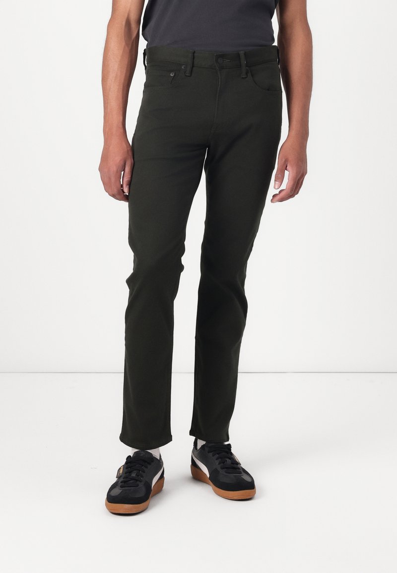 Pantalones ajustados de color verde oscuro, hechos de una tela suave, que cuentan con un diseño clásico de cinco bolsillos y cierre de botón metálico. Combinados con zapatillas negras.