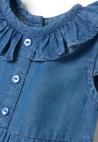 Vestido de mezclilla con cuello con volantes, botones azules y mangas cortas. La tela tiene una textura suave y un color azul uniforme.