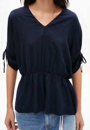 Woman wearing a navy blue V-neck blouse with cinched waist and tied short sleeves paired with light blue jeans.