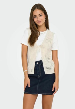 Knit cream cardigan with openwork design, tied front, paired with a white t-shirt and dark denim skirt, featuring a button and pockets.