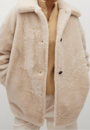 Beige plysset oversized jakke med stor krave over en kamelfarvet ribstrikket lynlås-sweater og lysfarvede bukser.