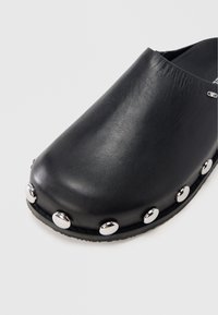 Black leather clog with rounded toe, featuring silver metallic studs along the base and smooth texture. No heel, slip-on design.