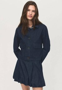 Temna denim jakna z dvema sprednjima žepoma, s kragnatim ovratnikom in gumbom za zapiranje, v kombinaciji z razširjenim denim krilom.
