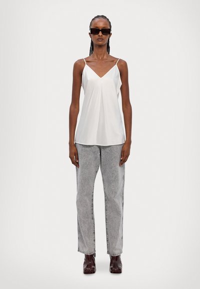 Top blanco sin mangas con diseño de escote en V; ajuste suelto. Combinado con jeans de cintura alta en gris claro y botas de tobillo marrón oscuro.