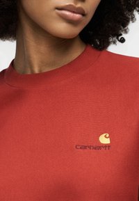 Γυναίκα που φοράει κόκκινο φούτερ με στρογγυλή λαιμόκοψη και κίτρινο λογότυπο Carhartt κεντημένο στο στήθος.