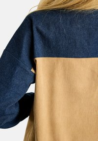 Farmering ing kék felső résszel és bordázott bézs alsó résszel. Laza ujjakkal és kontrasztos textúrákkal rendelkezik.