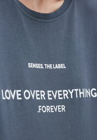 Nahaufnahme eines blauen Shirts mit weißem Text, der "SENSES. THE LABEL" und "LOVE OVER EVERYTHING .FOREVER" auf der Vorderseite liest.