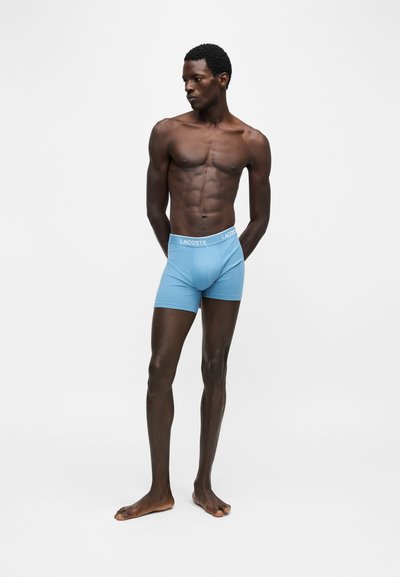 Homme debout, regardant à gauche, portant un boxer bleu clair avec ceinture "LACOSTE", mains derrière le dos, fond uni.