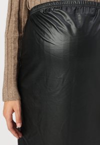 Jupe en simili cuir noir et brillant avec une taille élastique. Le gros plan montre la texture lisse et la finition élégante, associée à un haut texturé marron.