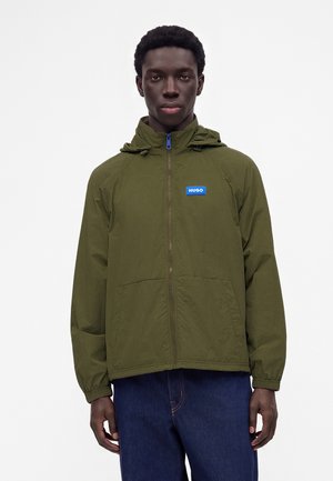 Jeune homme portant une veste zippée vert olive avec un patch logo bleu "HUGO" et un jean bleu foncé, debout devant un fond uni.