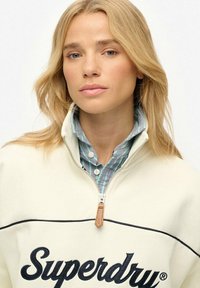 Cream-colored half-zip sweatshirt with navy "Superdry" logo, featuring a textured fabric and a tan leather zipper pull, layered over a blue and green striped shirt.