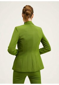 Femme aux cheveux attachés portant un blazer ajusté vert vif et un pantalon assorti, debout, les mains sur les hanches, devant un fond uni.