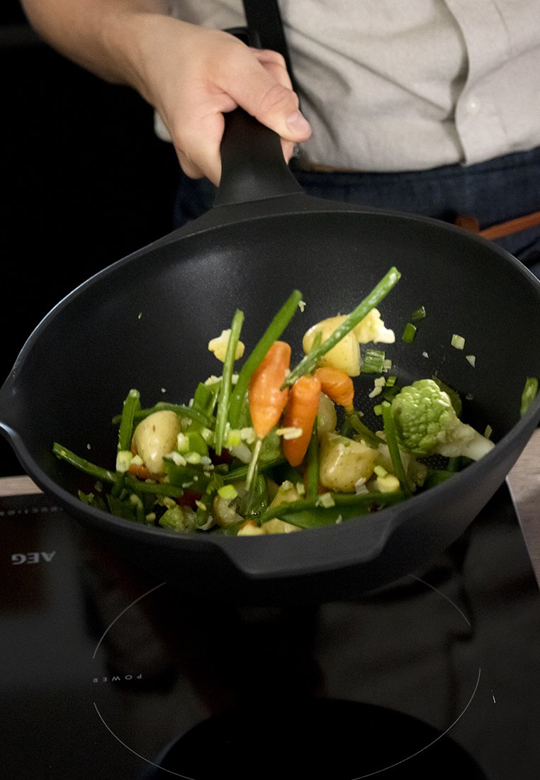 Hand hält eine schwarze Bratpfanne und wirft gehacktes Gemüse, darunter Karotten, grüne Bohnen und Kartoffeln, über einem Induktionsherd in die Luft.