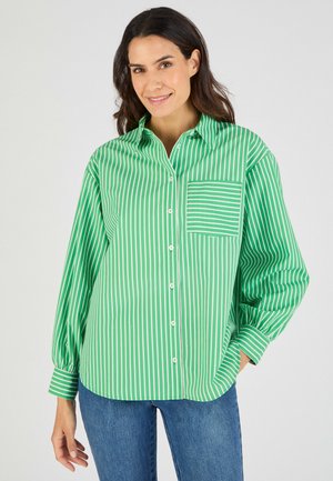 Mujer sonriente con una camisa verde y blanca a rayas verticales, abotonada, con un bolsillo delantero y vaqueros azules, sobre un fondo blanco.
