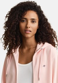 Kobieta z kręconymi włosami, ubrana w jasnoróżową bluzę z kapturem na suwak, na białym prążkowanym topie i cienkim złotym naszyjnikiem, zwrócona przodem.