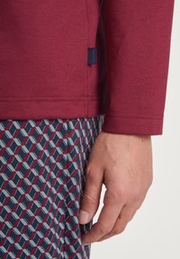 Chemise à manches longues bordeaux avec texture côtelée, étiquette sur le côté. Pantalon bleu marine à motifs avec des formes géométriques rouges et blanches. Main partiellement visible.